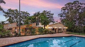 A swimming pool with a tiled edge, surrounded by a fence and trees. There are buildings with a brick facade and a covered area with lights on. - Serendipity Gardens! (Joyner)