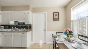 A kitchen with white cabinets, a tiled backsplash, and a dining area with a table set for four. - Grill, Relax, or Play at Home Away From Home (Shreveport)