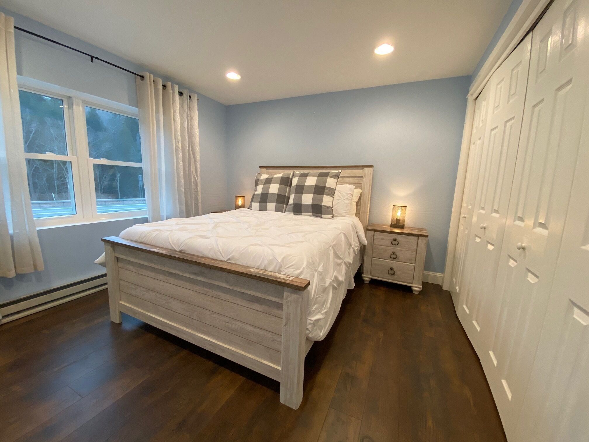 A bedroom with a wooden bed, a nightstand with a lamp, a window with curtains, and a wardrobe.