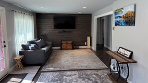A modern living room with a grey sofa, a wooden coffee table, a large flat-screen TV mounted on a dark wooden wall, and a framed quote on the wall. - Newly Renovated AC 4 ACRES 3000sqft home with Full Game Room (Hunter)