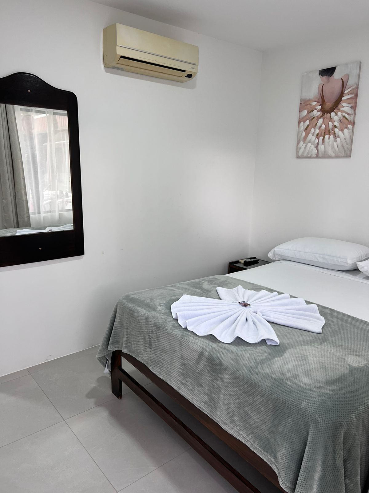 A hotel room with a bed, a window with curtains, an air conditioner, and a painting on the wall.