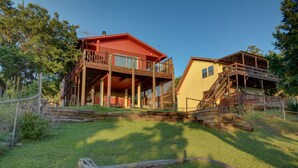A rustic cabin with a red roof and wooden deck, surrounded by a grassy area and trees. - Amazing Sunset View on Canyon Lake! (Canyon Lake)