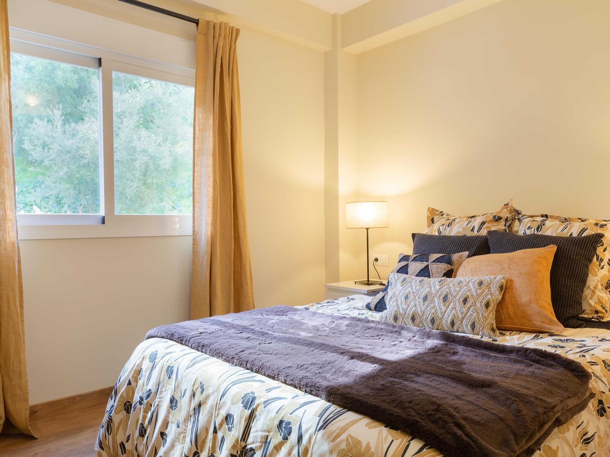 Un dormitorio con cama, mesita de noche, lámpara y ventana con cortinas.