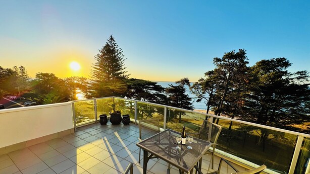 A balcony with a table and chairs, overlooking a scenic view of trees and a body of water during sunset. - STYLISH ISLAND GETAWAY - BEACHFRONT APARTMENT - COWES - Phillip Island (Cowes)