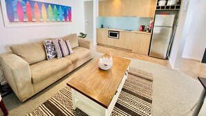 A living room with a beige sofa, a wooden coffee table, and patterned rugs. A kitchen is visible in the background with wooden cabinets, a microwave, and a refrigerator. - STYLISH ISLAND GETAWAY - BEACHFRONT APARTMENT - COWES - Phillip Island (Cowes)