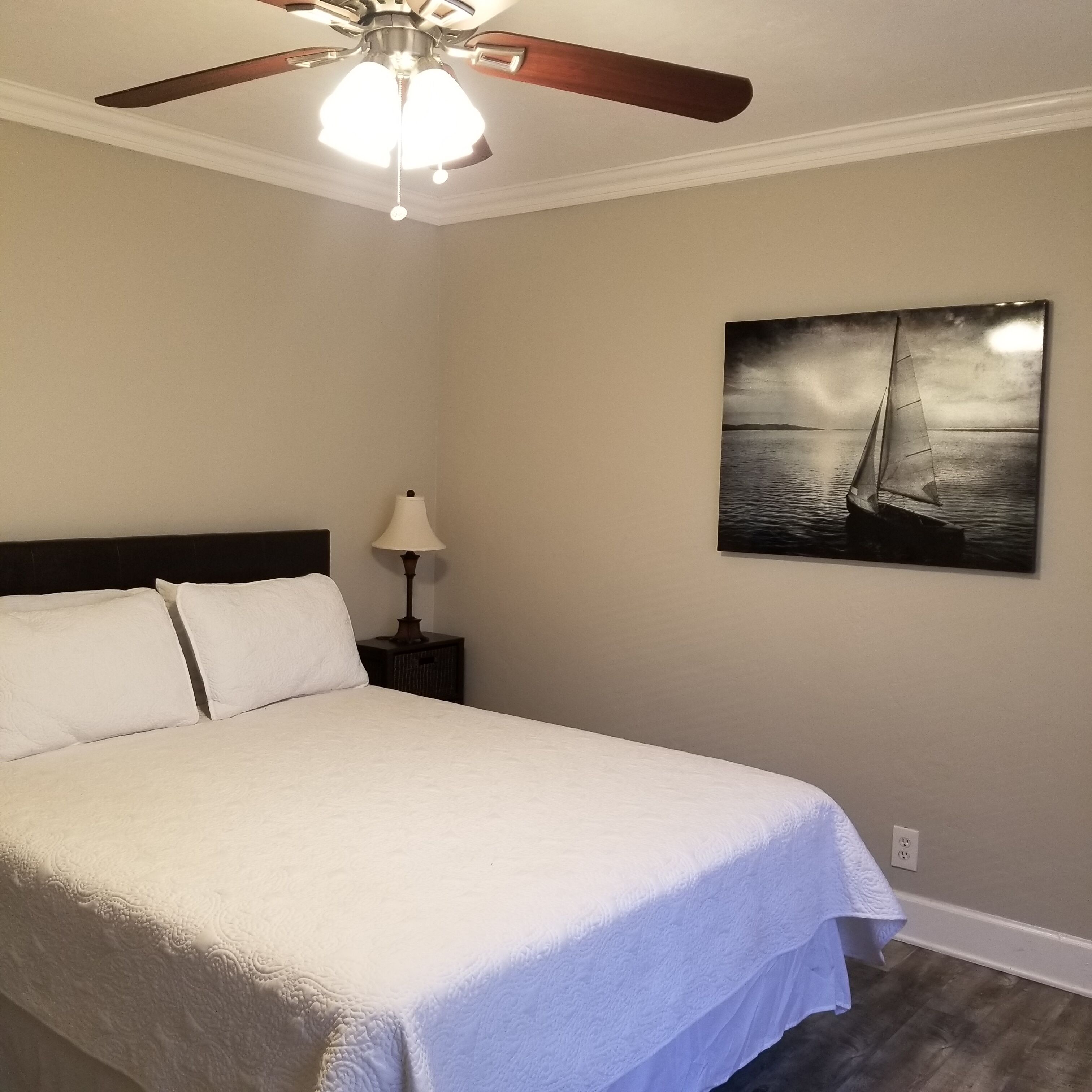 Een slaapkamer met een bed, een plafondventilator, een nachtkastje met een lamp en een schilderij van een zeilboot aan de muur.
