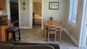 Una sala de estar con un sofá marrón oscuro, un comedor con mesa y sillas de madera, una cocina con encimera oscura y armarios de madera, y un pasillo que conduce a un dormitorio con cama y puerta.