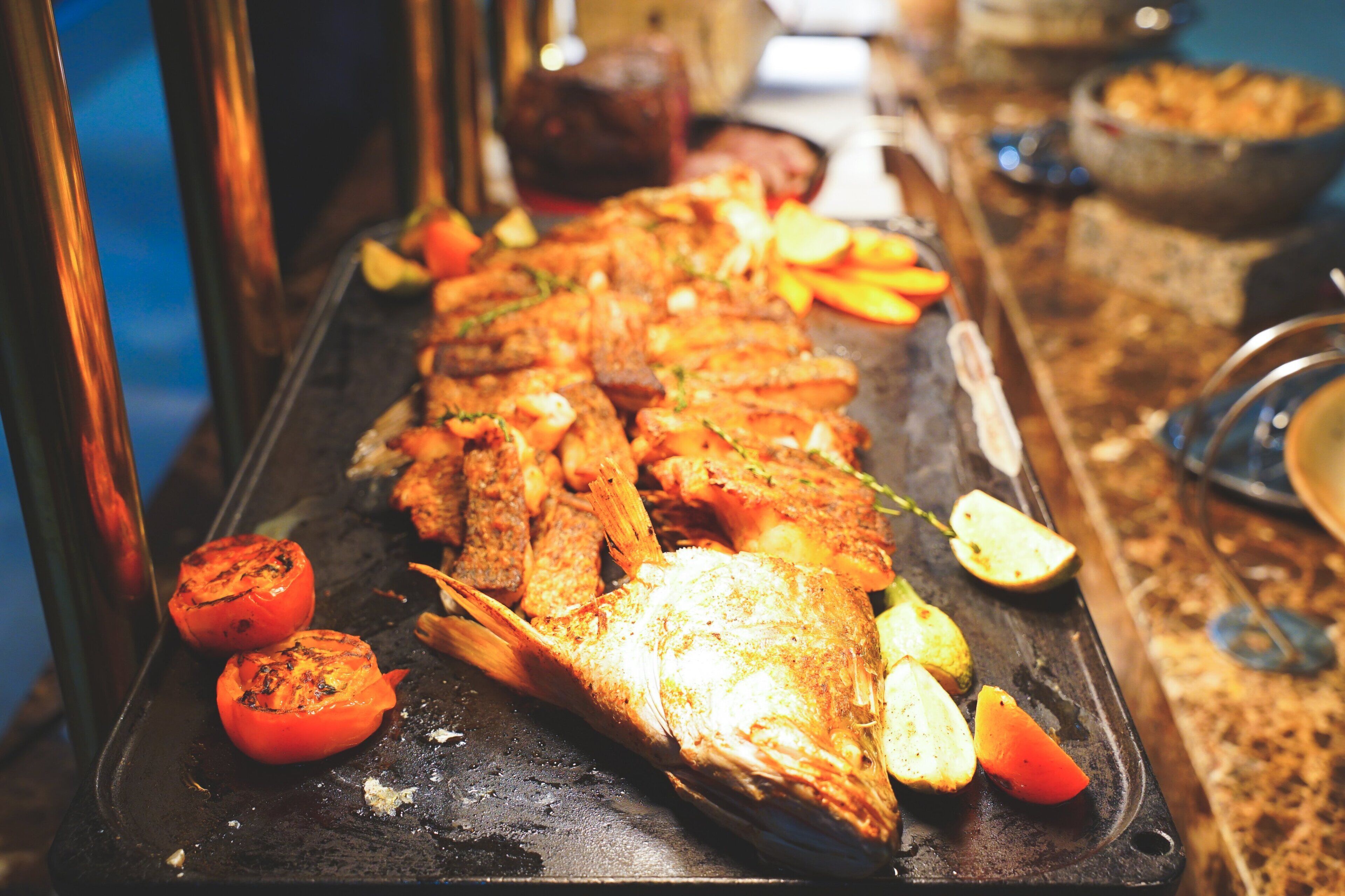 A long fish grill with grilled vegetables and a variety of dishes in the background.