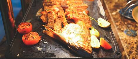 A long fish grill with grilled vegetables and a variety of dishes in the background.
