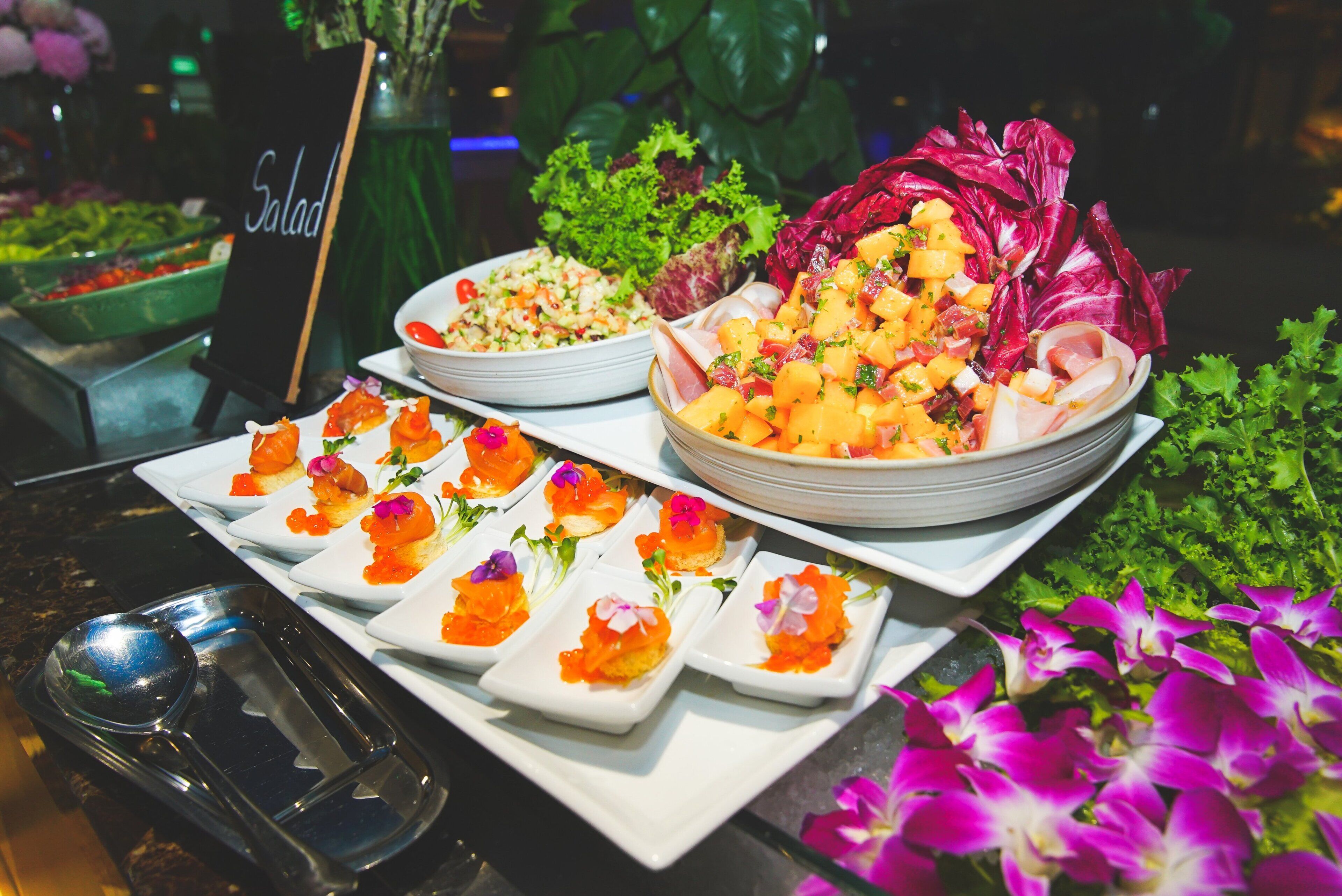 A buffet with various dishes including salads, a bowl of mango chunks, and small plates with salmon garnished with flowers.