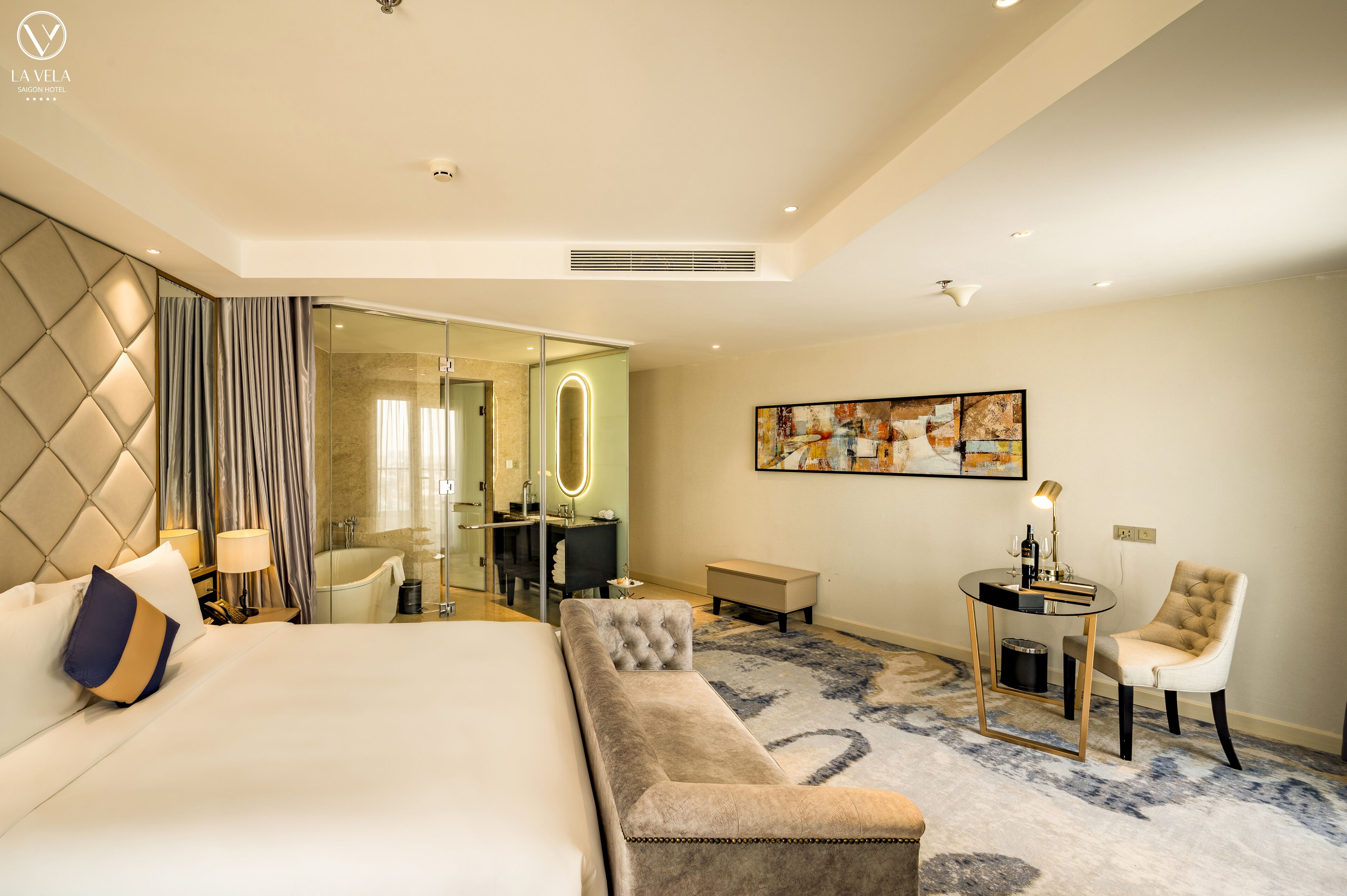 A modern hotel room with a large bed, a seating area, a desk, and a bathroom with a glass shower enclosure.