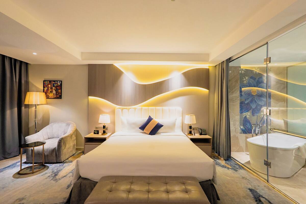 Una habitación de hotel moderna con una cama grande, una zona de estar con una silla y una mesita, un baño con paredes de vidrio e iluminación ambiental.