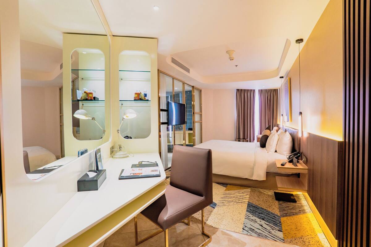 Habitación de hotel con cama, escritorio con silla, espejo, televisor y mesita de noche con lámpara.