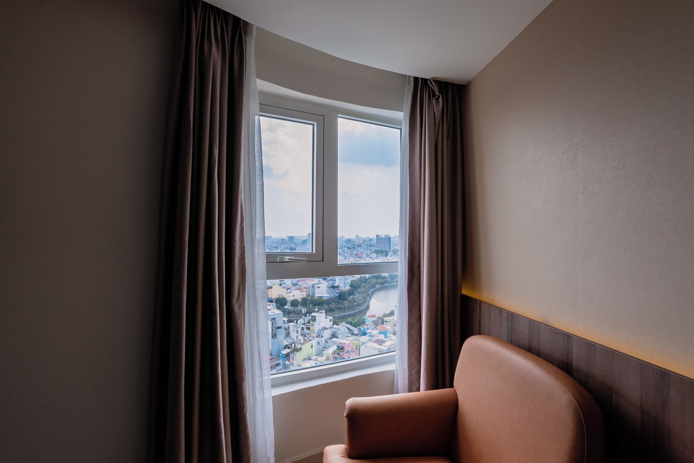 Una habitación con un amplio ventanal, vista a un paisaje urbano y un sillón de cuero marrón.