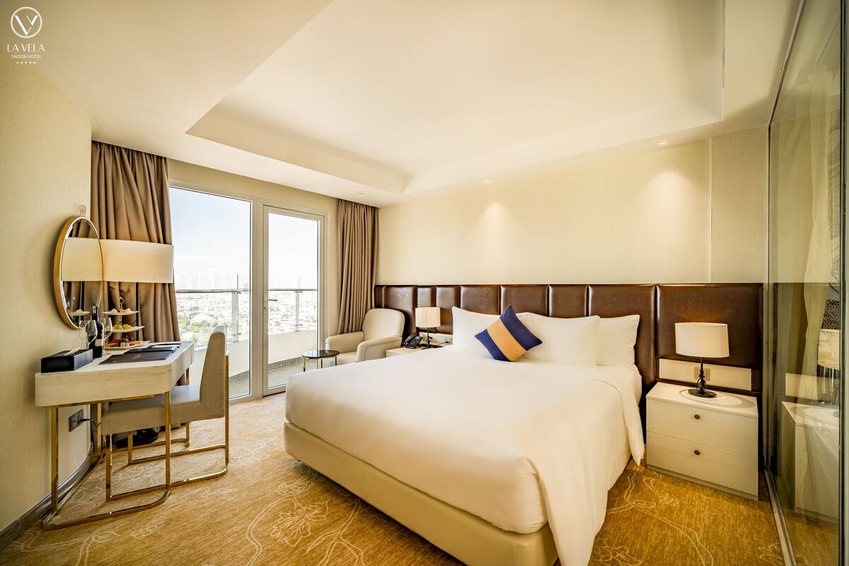 Habitación de hotel con una cama grande, un escritorio con silla, una mesita de noche y un ventanal con cortinas.