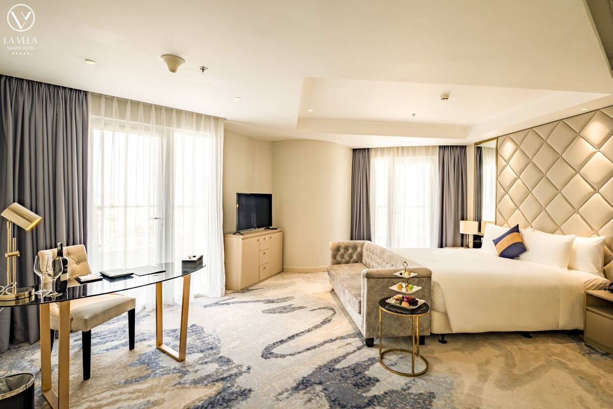 Habitación de hotel con una cama grande, un sofá, un escritorio con computadora, un televisor y una mesa de comedor con lámpara.
