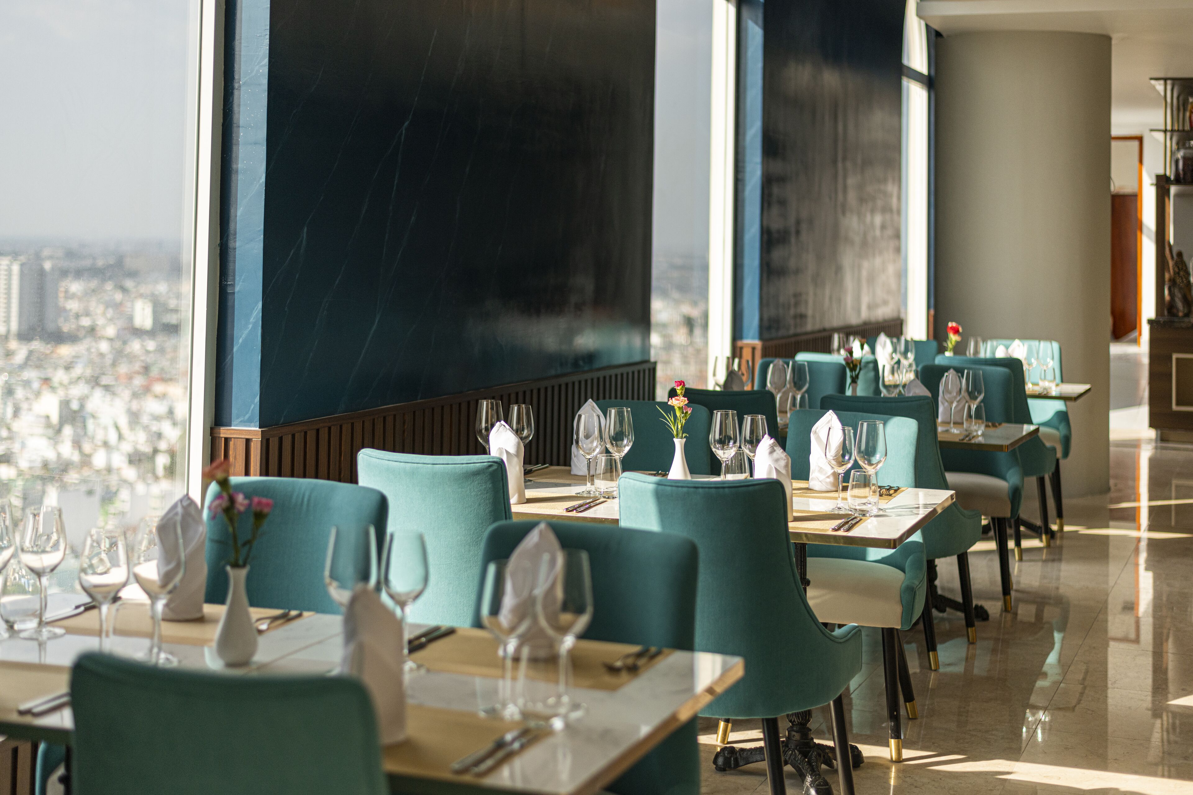 En restaurang med turkosa stolar och bord, stora fönster med utsikt över staden och en modern inredning.