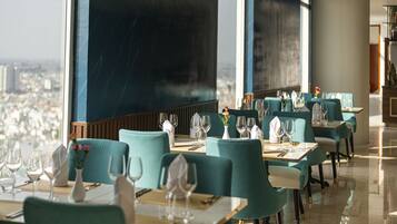 En restaurang med turkosa stolar och bord, stora fönster med utsikt över staden och en modern inredning.