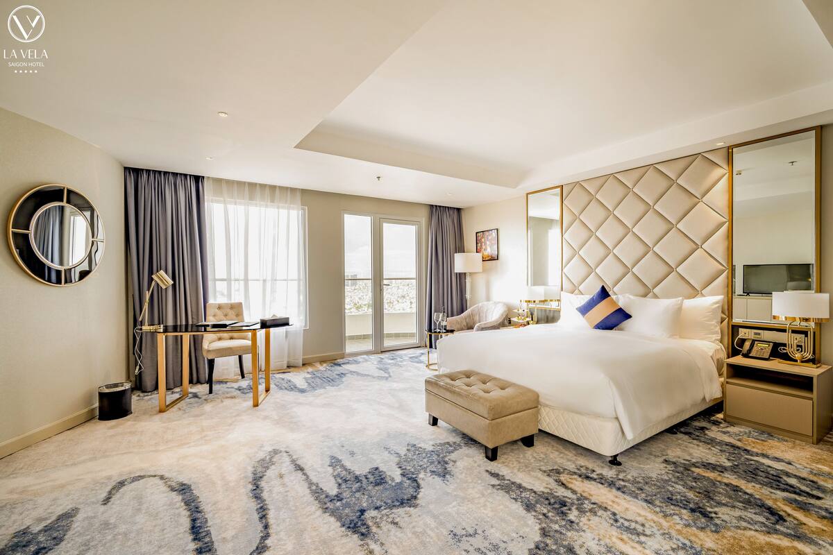 Habitación de hotel con una cama grande, un escritorio, una silla, un pequeño reposapiés, un espejo redondo y un ventanal con cortinas.