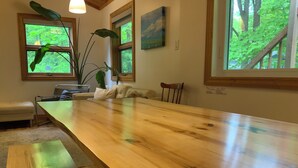 Una larga mesa de comedor de madera con bancos, una silla y una planta en una habitación con ventanas amplias y un cuadro en la pared.