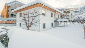 Una casa moderna de dos pisos con techo nevado, un muelle de madera y vistas a montañas al fondo.
