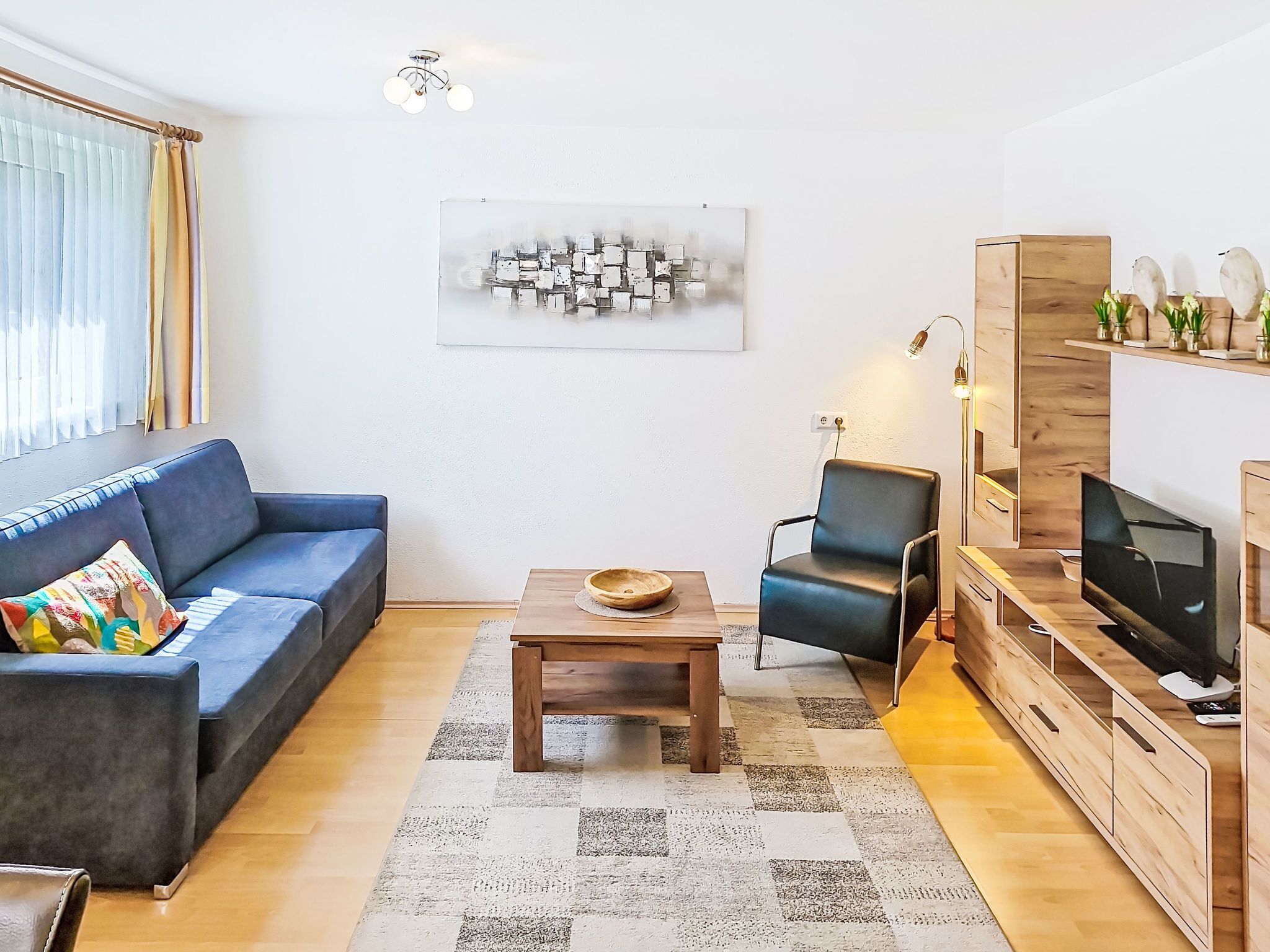 Ein modernes Wohnzimmer mit einem blauen Sofa, einem Holztisch, einem schwarzen Sessel, einem Fernseher und einer Wanddekoration.