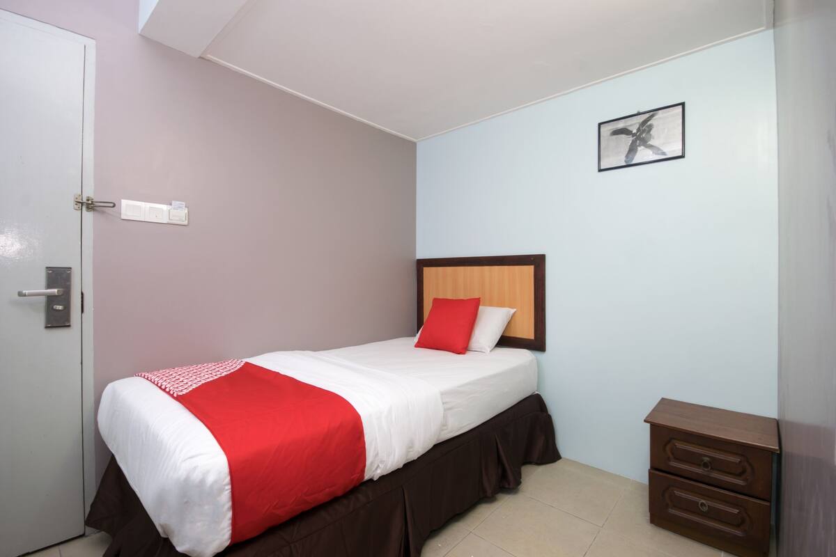 A single bed with a wooden headboard, a red and white bedspread, and a small wooden nightstand with drawers.