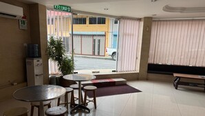 A hotel lobby with a reception desk, a water dispenser, a potted plant, a seating area with a bench and stools, and a large window with blinds. - Hotel O Hotel Oriental (Kota Kinabalu)