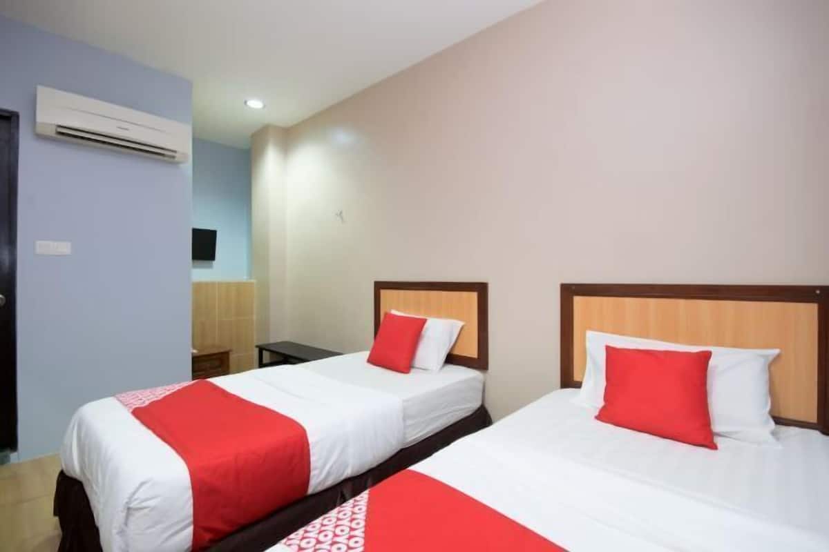Two single beds with red and white bedding, a wall-mounted air conditioner, and a small table with a lamp.