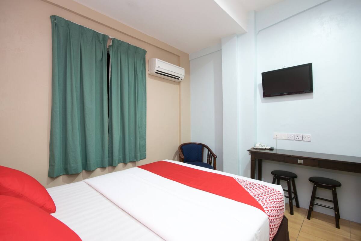 A hotel room with a bed, green curtains, an air conditioning unit, a television, a desk with two stools, and a chair.