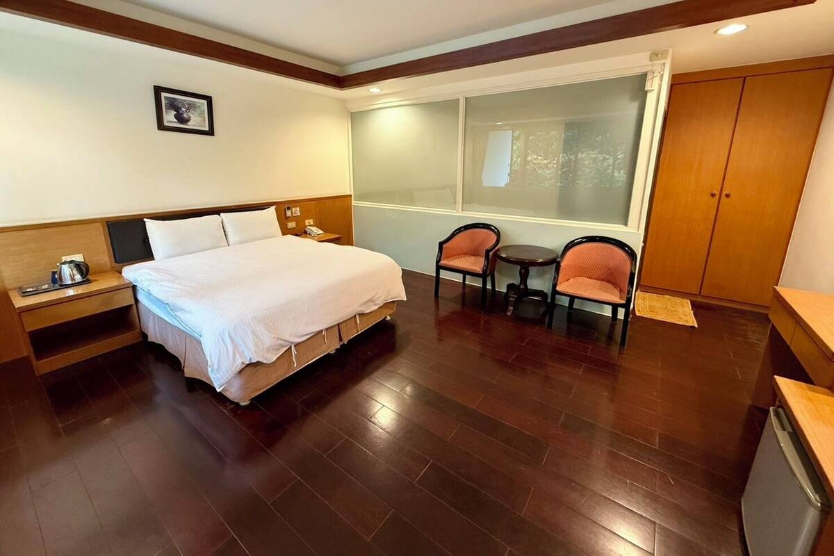 A hotel room with a bed, bedside table, two chairs, a small table, a window, and a wardrobe.
