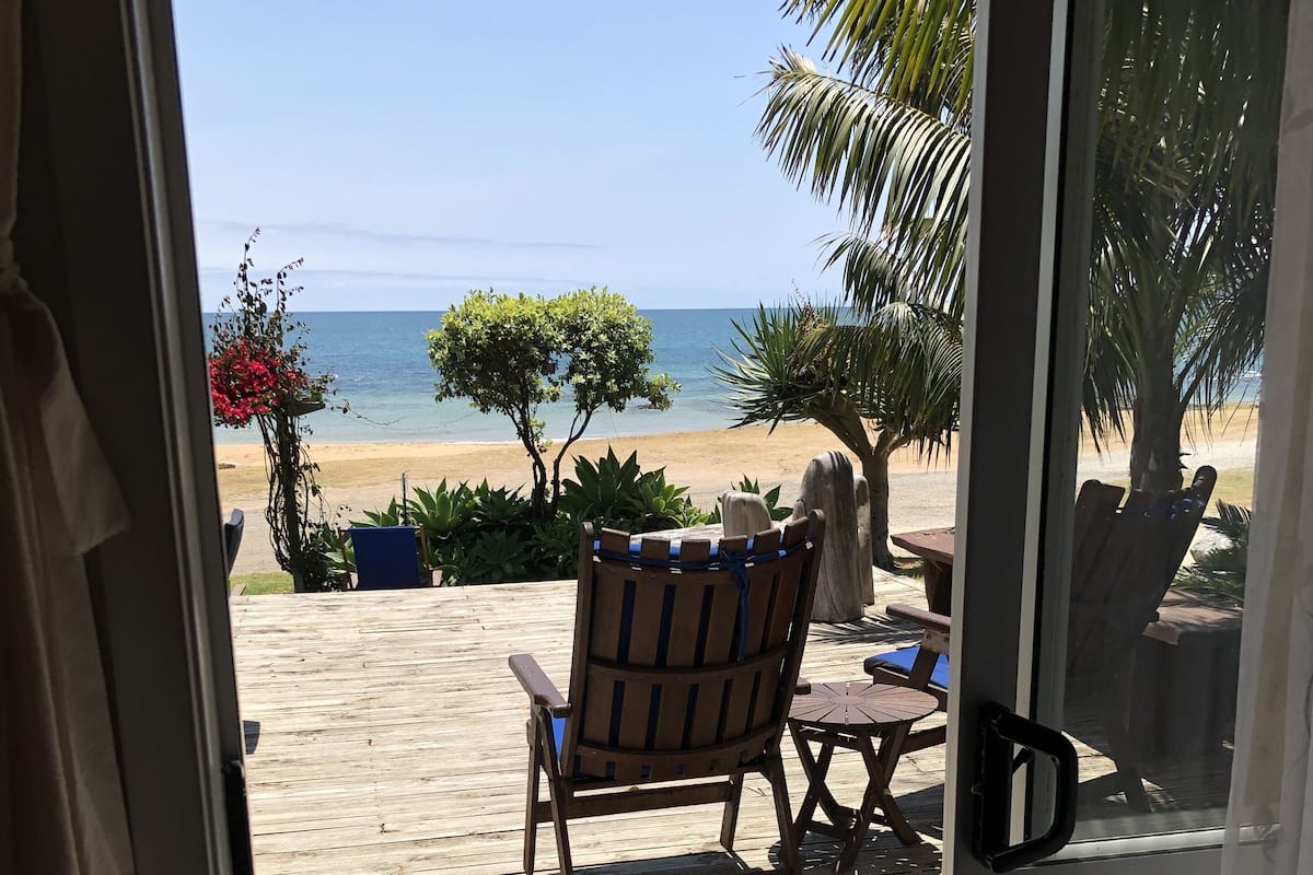 A wooden deck with a chair and table, overlooking a beach with palm trees and a clear sky.