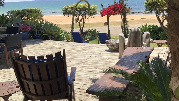 Una terraza de madera con mobiliario exterior, vista a la playa y plantas tropicales.