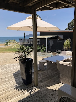 An outdoor patio with a view of the beach, a large umbrella, wicker furniture, and a potted plant. - Driftwood Lodge (Cable Bay)