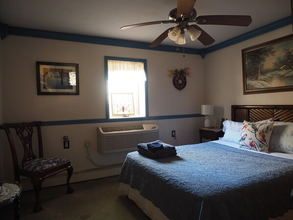 Une chambre à coucher avec un lit, une chaise, une fenêtre avec des rideaux, un ventilateur de plafond et un tableau accroché au mur.