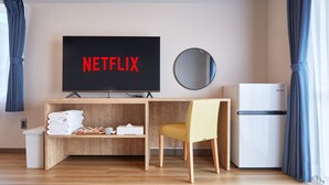 Une chambre d’hôtel moderne avec un bureau en bois, un téléviseur affichant « NETFLIX », un miroir rond, une chaise jaune, un réfrigérateur blanc et une petite poubelle.