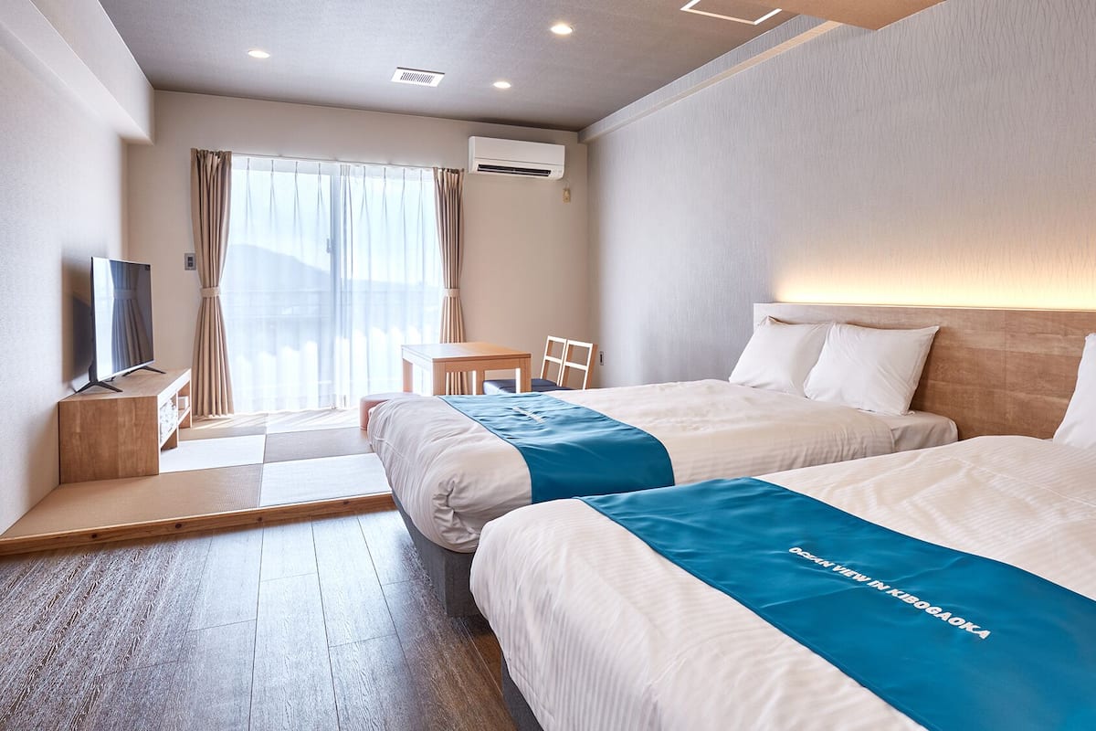 A hotel room with two beds, a TV, a desk, and a view of the outdoors.