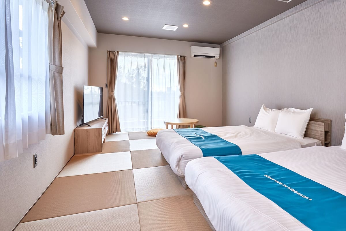 A hotel room with two beds, a TV, a small table, and an air conditioning unit.