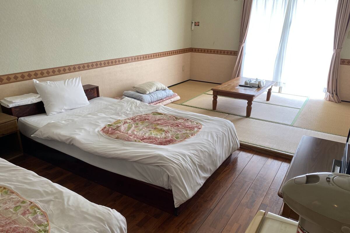 A hotel room with two beds, a small table, a chair, a wooden floor, and a window with curtains.