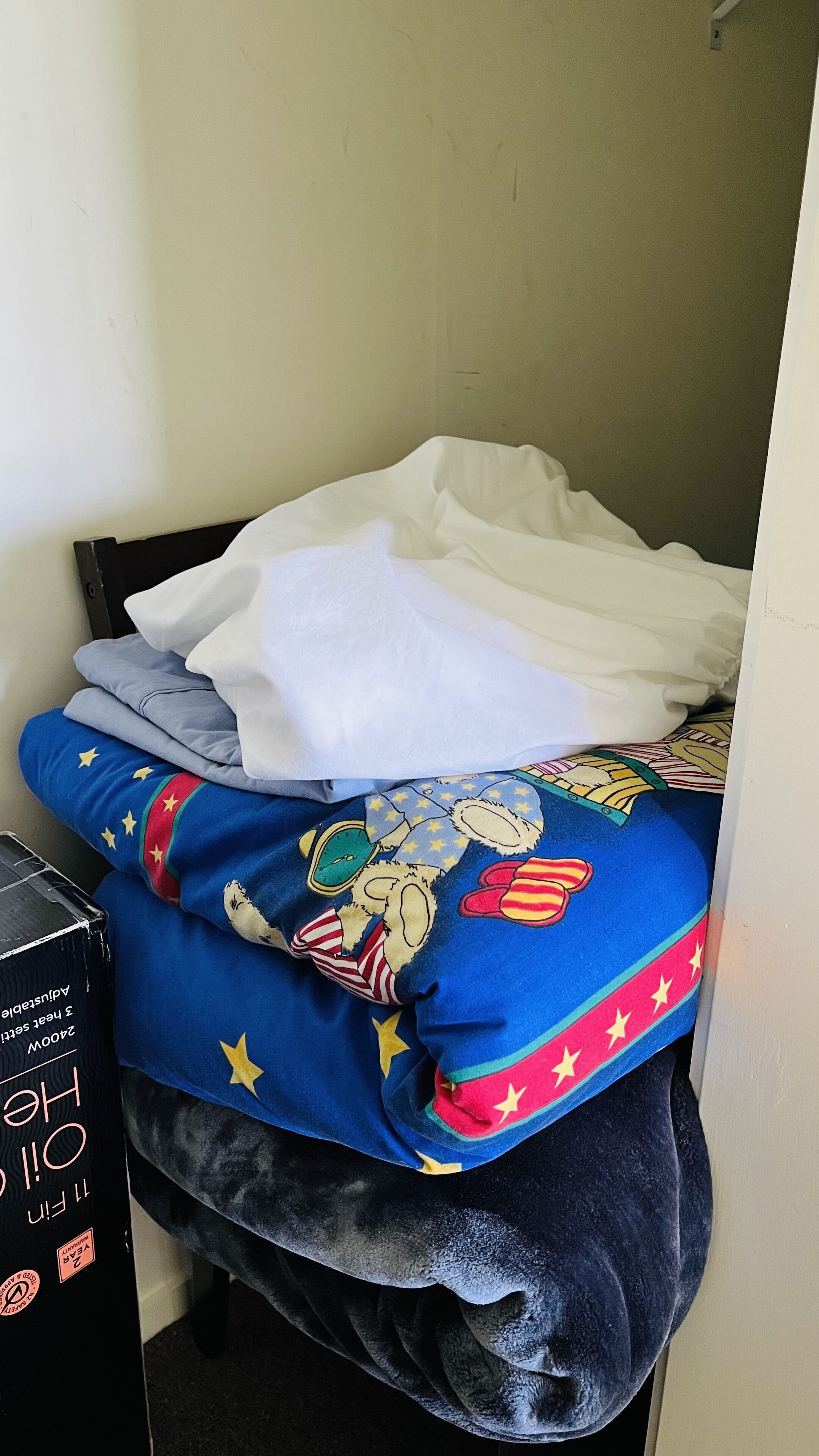 Una cama con sábana blanca, manta azul con diseño de superhéroe y una caja negra en el suelo.