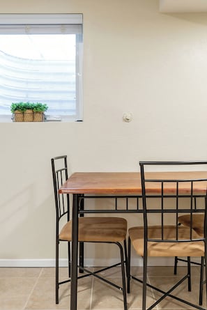 A dining area with a wooden table and metal chairs, a window with potted plants, and a white wall.