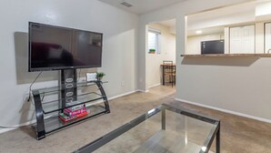 Una sala de estar con un televisor de pantalla plana sobre un pedestal de vidrio, una mesa de centro de vidrio y piso alfombrado.