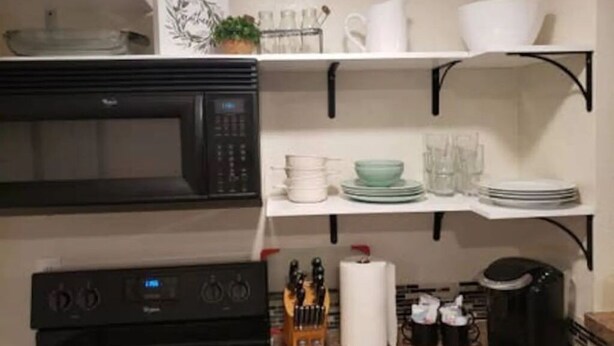 Una cocina con microondas, horno y cafetera. Hay estantes con vajilla y una planta.