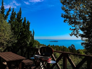 A wooden deck with a barbecue grill and a table, overlooking a clear blue sea and a distant island.