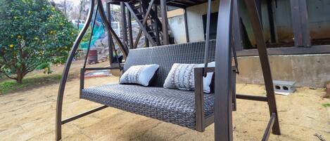 A metal swing chair with cushions, set outdoors on a sandy surface, with a building and a tree in the background.