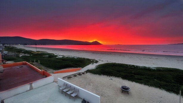 A beach with a sunset, a sandy area with a table and chairs, and a building in the foreground.