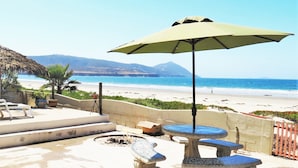 A beachside patio with a table and chairs, a thatched umbrella, and a view of the beach and ocean.