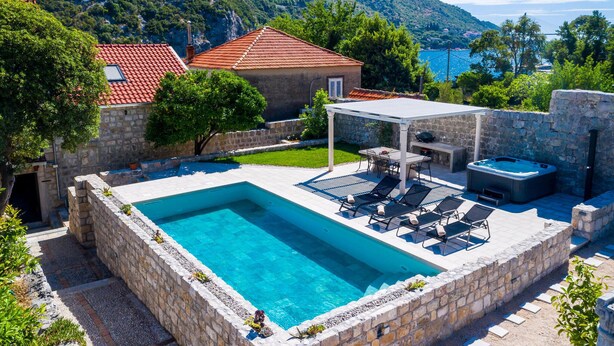 A rectangular swimming pool with a stone border, a covered patio area with outdoor furniture, and a view of a mountainous landscape in the background.