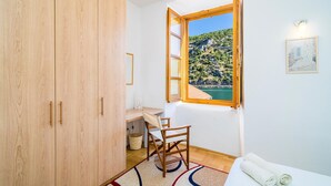 A room with a wooden wardrobe, a desk with a chair, a window with a view of a coastal landscape, and a bed with a rolled-up towel.
