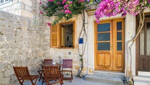 A stone building with a wooden door and window shutters, a table with chairs, and pink bougainvillea flowers.
